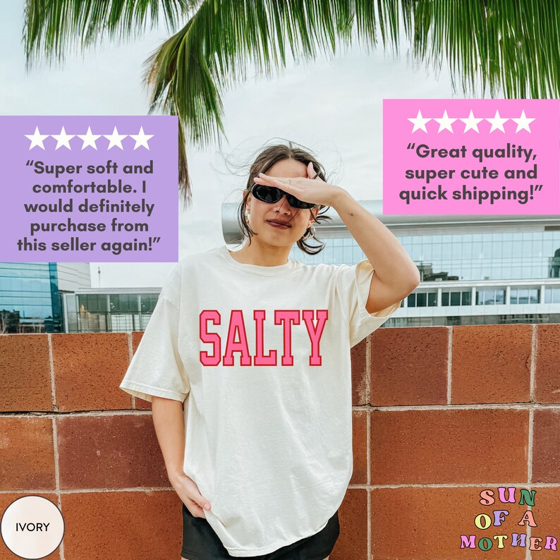 May include: A white t-shirt with the word "SALTY" printed in pink letters. The shirt is being worn by a person with sunglasses. The background is a brick wall with a palm tree and a building in the distance.
