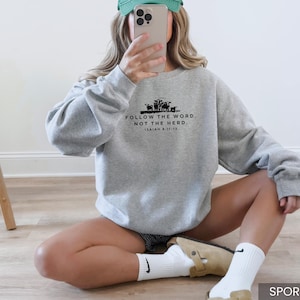 May include: Sport grey sweatshirt with the text "FOLLOW THE WORD, NOT THE HERD" and Isaiah 8:11-13. The sweatshirt has a crew neck and long sleeves. The image also shows a wooden stool.