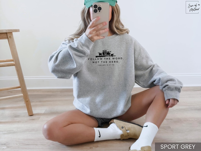 May include: Sport grey sweatshirt with the text "FOLLOW THE WORD, NOT THE HERD" and Isaiah 8:11-13. The sweatshirt has a crew neck and long sleeves. The image also shows a wooden stool.