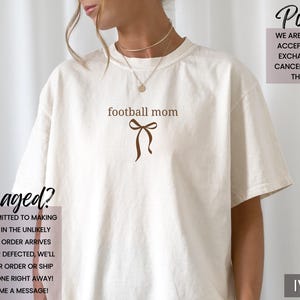 May include: Ivory-colored t-shirt with the text "football mom" in brown, along with a brown bow graphic. The shirt also features text regarding returns and damaged items. The brand logo "Sun of a Mother" is in the top left corner.
