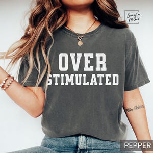 May include: A gray t-shirt with the text "OVER STIMULATED" printed in white on the front. The shirt is being worn by a person with long brown hair.