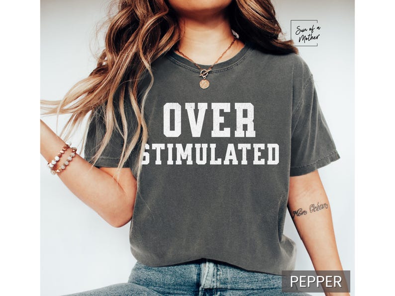 May include: A gray t-shirt with the text "OVER STIMULATED" printed in white on the front. The shirt is being worn by a person with long brown hair.