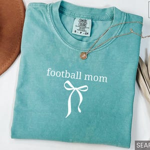 May include: Seafoam green t-shirt with the words "football mom" and a bow graphic in white. A gold necklace with a pendant rests on the shirt. The shirt is folded on a white surface.