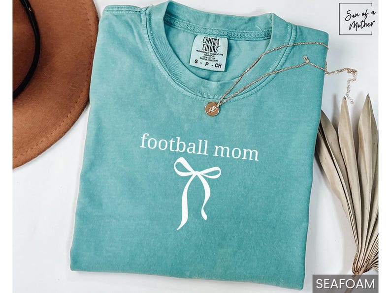 May include: Seafoam green t-shirt with the words "football mom" and a bow graphic in white. A gold necklace with a pendant rests on the shirt. The shirt is folded on a white surface.