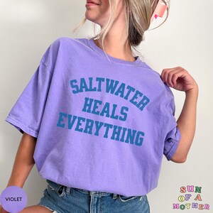 May include: A purple t-shirt with the text "Saltwater Heals Everything" in blue lettering. The shirt is being worn by a person with long blonde hair.