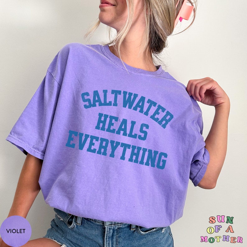 May include: A purple t-shirt with the text "Saltwater Heals Everything" in blue lettering. The shirt is being worn by a person with long blonde hair.