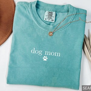 May include: A teal blue t-shirt with the text "dog mom" and a white paw print. The shirt is made of a soft, comfortable material.