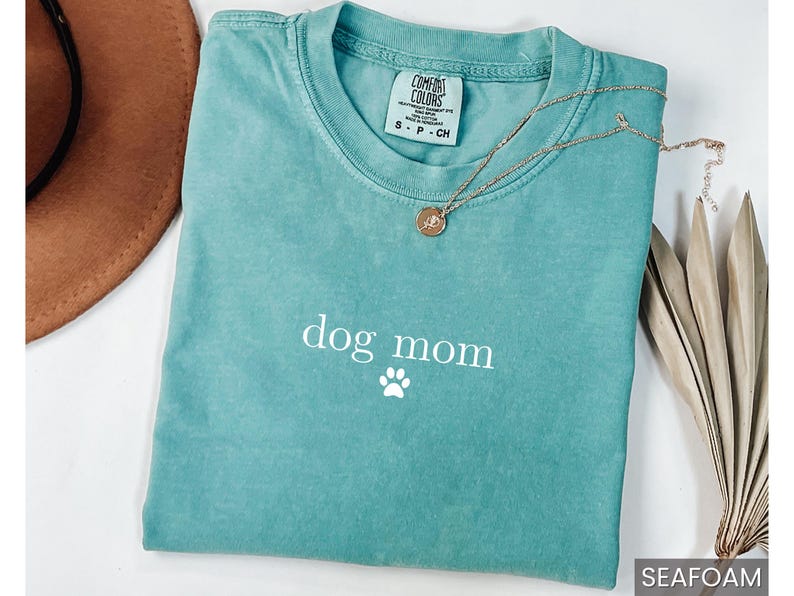 May include: A teal blue t-shirt with the text "dog mom" and a white paw print. The shirt is made of a soft, comfortable material.