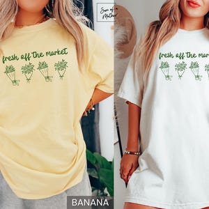 May include: A light yellow t-shirt and a white t-shirt with the text "fresh off the market" above a graphic of four bouquets. The shirts are casual and comfortable, suitable for everyday wear.