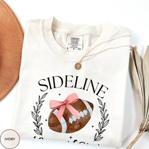 以下が含まれることがあります： アイボリー色のTシャツに、黒い文字で「SIDELINE」と書いてあり、その下にピンクのリボンが付いたフットボールの水彩画が描かれています。フットボールは葉のリースで囲まれています。