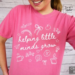 May include: Pink t-shirt with a white graphic design that says "helping little minds grow" and includes illustrations of school supplies like a pencil, apple, rainbow, and an A+.