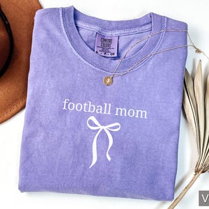 May include: A violet Comfort Colors t-shirt with the words "football mom" and a bow graphic in white. A gold necklace and a brown hat are also in the image. The shirt is folded and ready to wear.