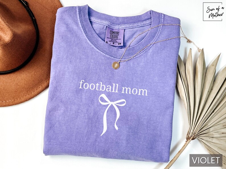 May include: A violet Comfort Colors t-shirt with the words "football mom" and a bow graphic in white. A gold necklace and a brown hat are also in the image. The shirt is folded and ready to wear.