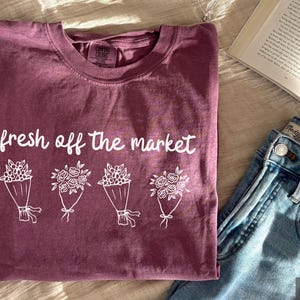 May include: Berry-colored t-shirt with the phrase "fresh off the market" and four bouquets of flowers in white. The shirt is paired with blue jeans and an open book.