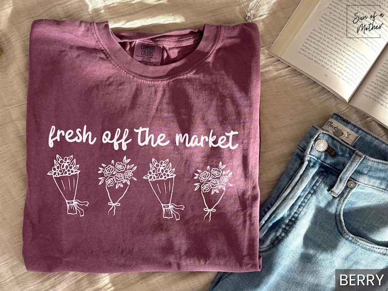 May include: Berry-colored t-shirt with the phrase "fresh off the market" and four bouquets of flowers in white. The shirt is paired with blue jeans and an open book.