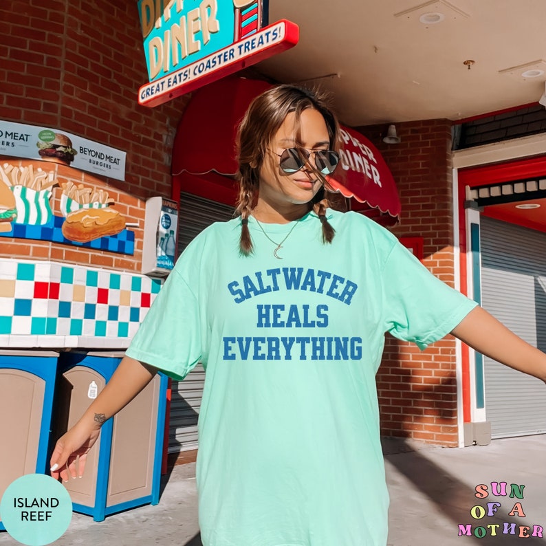 May include: A light green t-shirt with the text "Saltwater Heals Everything" in blue lettering. The shirt is worn by a person with long brown hair and sunglasses. The person is standing in front of a red and white building with a sign that says "Diner".