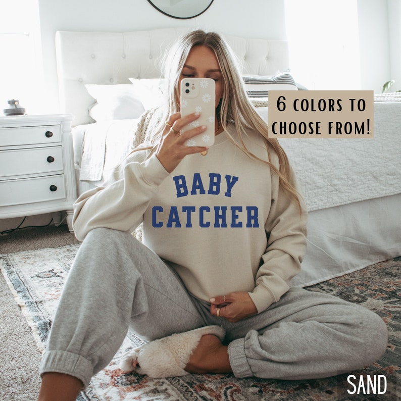 May include: A woman wearing a light beige sweatshirt with the text "BABY CATCHER" in blue lettering. The sweatshirt is a relaxed fit and has a crew neckline. The woman is sitting on a rug with her legs crossed.
