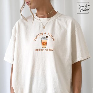 May include: Ivory-colored t-shirt with the text "feeling a little spicy today" in brown script. The graphic features a pumpkin spice latte with whipped cream and cinnamon sticks. The shirt is a relaxed fit.