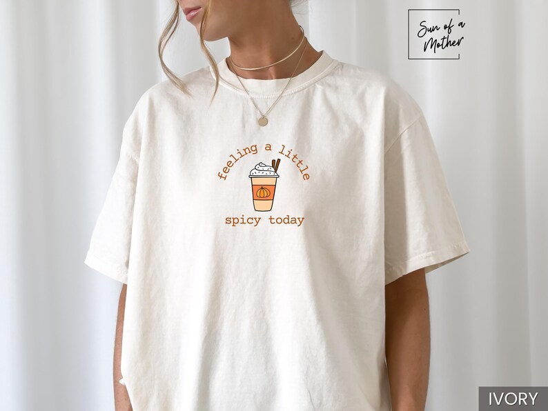 May include: Ivory-colored t-shirt with the text "feeling a little spicy today" in brown script. The graphic features a pumpkin spice latte with whipped cream and cinnamon sticks. The shirt is a relaxed fit.