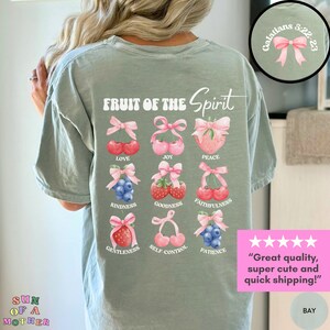 May include: A light green t-shirt with the text "FRUIT OF THE Spirit" printed on the back. The text is surrounded by nine images of fruit with pink bows, each representing a different fruit of the spirit: love, joy, peace, kindness, goodness, faithfulness, gentleness, self-control, and patience. The text "Galatians 5:22-23" is printed in a smaller font on the top right corner of the shirt.