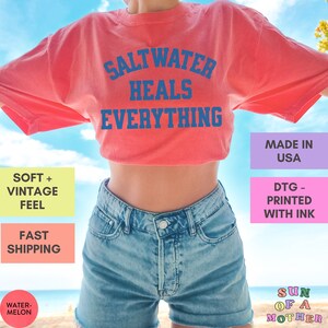 May include: A red t-shirt with the text "Saltwater Heals Everything" in blue. The shirt is being worn by a person with blue denim shorts. The text "Made in USA" is in a purple box and the text "DTG - Printed with Ink" is in a pink box.