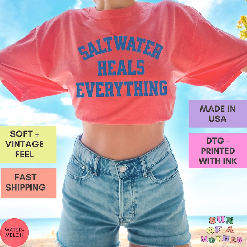 May include: A red t-shirt with the text "Saltwater Heals Everything" in blue. The shirt is being worn by a person with blue denim shorts. The text "Made in USA" is in a purple box and the text "DTG - Printed with Ink" is in a pink box.