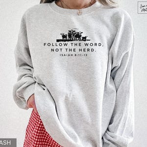 May include: Light gray sweatshirt with the text "FOLLOW THE WORD, NOT THE HERD." and "ISAIAH 8:11-13" in black. A black silhouette of cows is above the text. The sweatshirt is long-sleeved and has a crew neck.