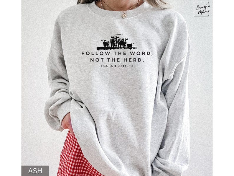 May include: Light gray sweatshirt with the text "FOLLOW THE WORD, NOT THE HERD." and "ISAIAH 8:11-13" in black. A black silhouette of cows is above the text. The sweatshirt is long-sleeved and has a crew neck.