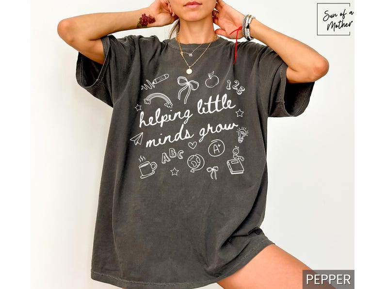 May include: A dark gray t-shirt with a white graphic design that says "helping little minds grow" and features various school-related icons. The shirt is a relaxed fit and has a crew neck.