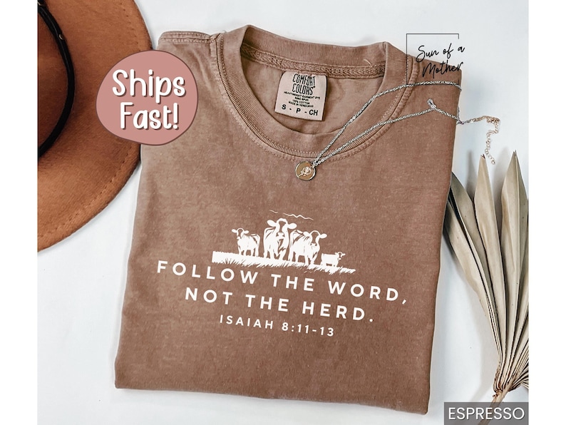 Peut inclure: Un t-shirt marron avec un graphique blanc de vaches et le texte "FOLLOW THE WORD, NOT THE HERD. ISAIAH 8:11-13".