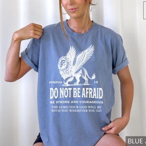 May include: Blue jean-colored t-shirt featuring a white graphic of a winged lion and the text "DO NOT BE AFRAID." The shirt also includes the Bible verse Joshua 1:9. The shirt is a comfortable, casual style.