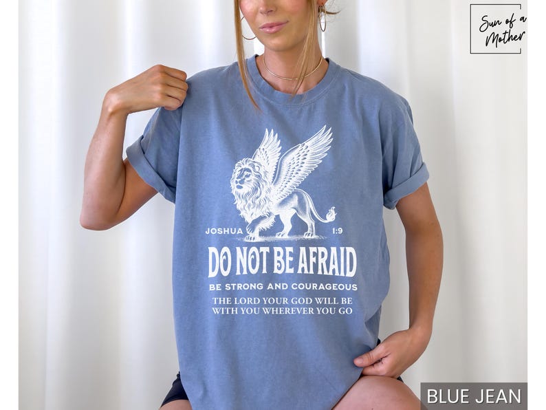 May include: Blue jean-colored t-shirt featuring a white graphic of a winged lion and the text "DO NOT BE AFRAID." The shirt also includes the Bible verse Joshua 1:9. The shirt is a comfortable, casual style.