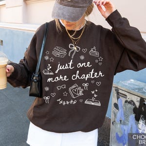 May include: Chocolate brown sweatshirt with the text "just one more chapter" in white. The design includes books, hearts, stars, glasses, and bows. The sweatshirt is a casual style.