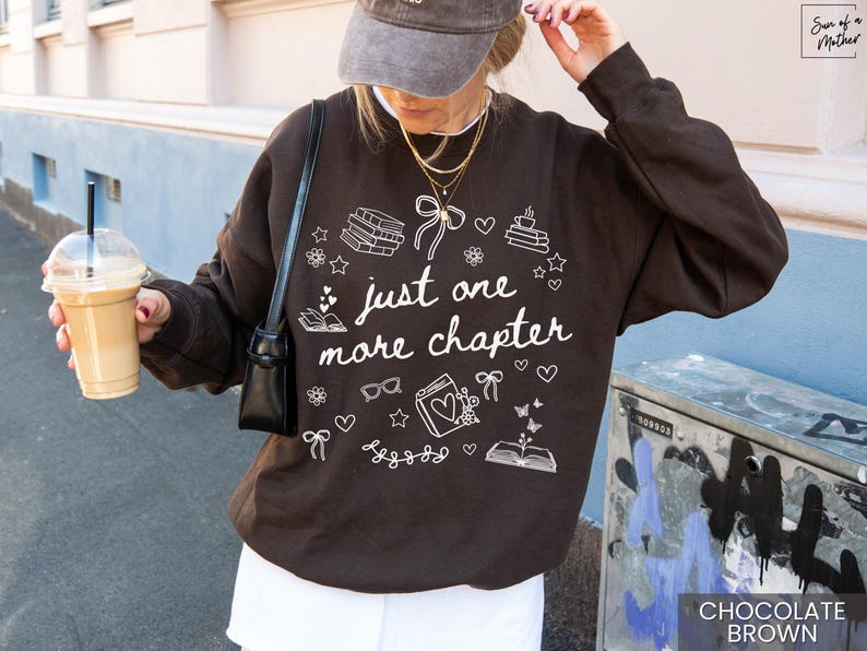 May include: Chocolate brown sweatshirt with the text "just one more chapter" in white. The design includes books, hearts, stars, glasses, and bows. The sweatshirt is a casual style.