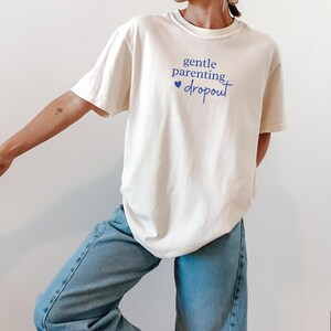 Puede incluir: Camiseta de color marfil con el texto "gentle parenting dropout" en escritura azul. La camiseta se combina con vaqueros azul claro. La imagen est&aacute; tomada frente a un fondo blanco. La marca "Sun of a Mother" est&aacute; en la esquina inferior izquierda.
