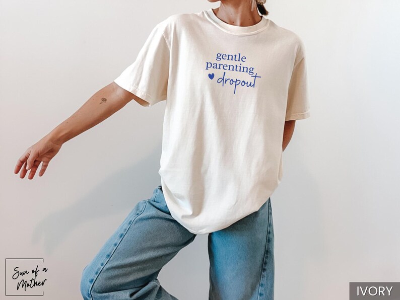 Puede incluir: Camiseta de color marfil con el texto "gentle parenting dropout" en escritura azul. La camiseta se combina con vaqueros azul claro. La imagen est&aacute; tomada frente a un fondo blanco. La marca "Sun of a Mother" est&aacute; en la esquina inferior izquierda.