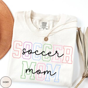 May include: Ivory t-shirt with the text "SOCCER mom" in rainbow colors with black outlines. The text is in a varsity style font.