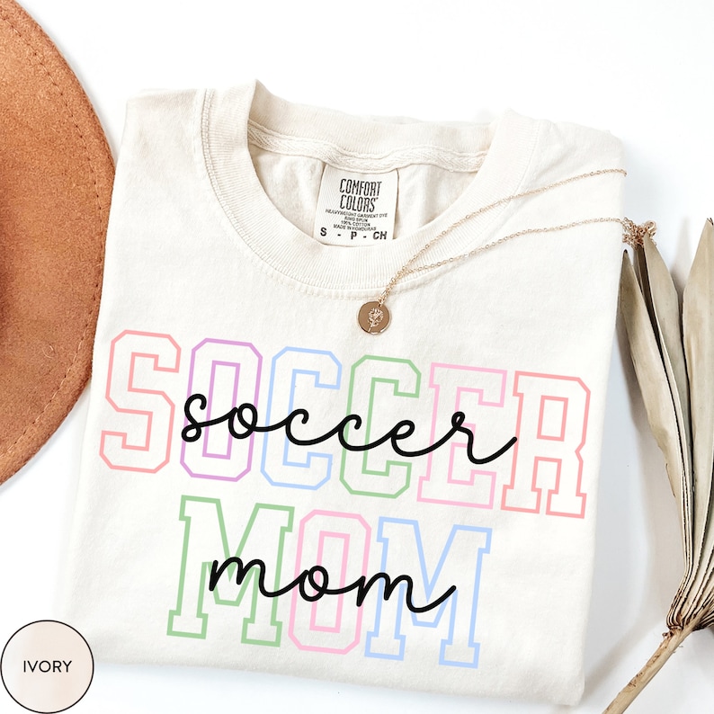 May include: Ivory t-shirt with the text "SOCCER mom" in rainbow colors with black outlines. The text is in a varsity style font.