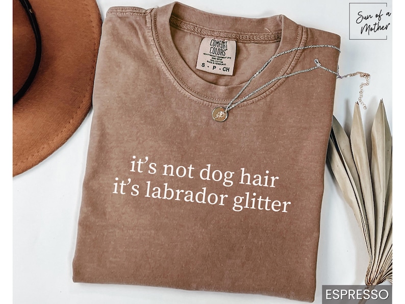 May include: Espresso-colored t-shirt with the text "it's not dog hair it's labrador glitter" in white. A silver necklace with a pendant rests on the shirt. A brown hat is partially visible.