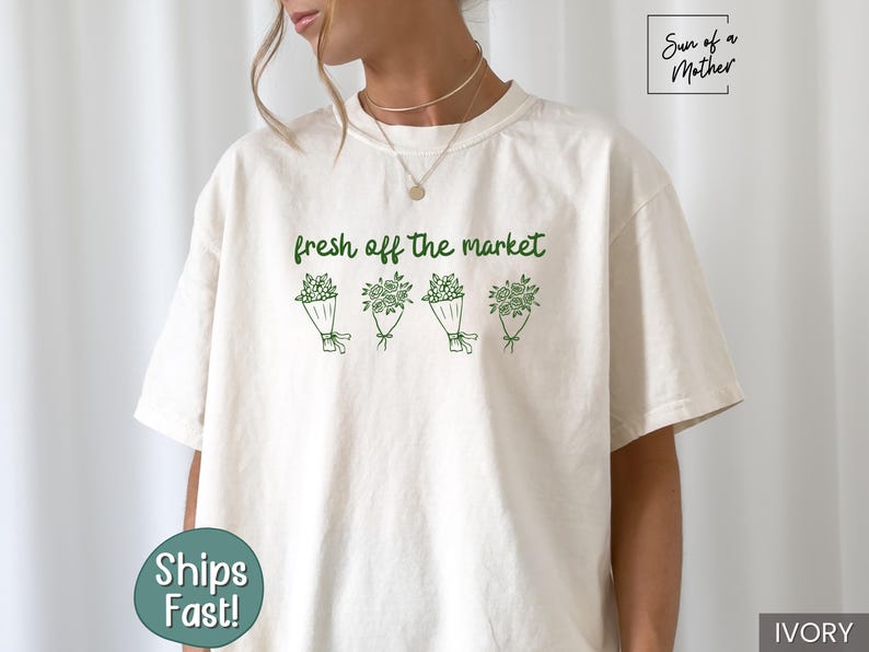 May include: Ivory-colored t-shirt with the text "fresh off the market" above four green floral bouquets. The shirt has a relaxed fit and short sleeves. The brand name "Sun of a Mother" is in the upper right corner.