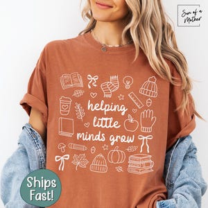 May include: Rust-colored t-shirt with the phrase "helping little minds grow" in white, surrounded by white illustrations of books, a coffee cup, and other school-related items. The shirt is worn by a person.