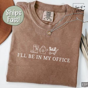 May include: A brown t-shirt with the text "I'll be in my office" and a graphic of gardening tools, including boots, a watering can, and a wheelbarrow.