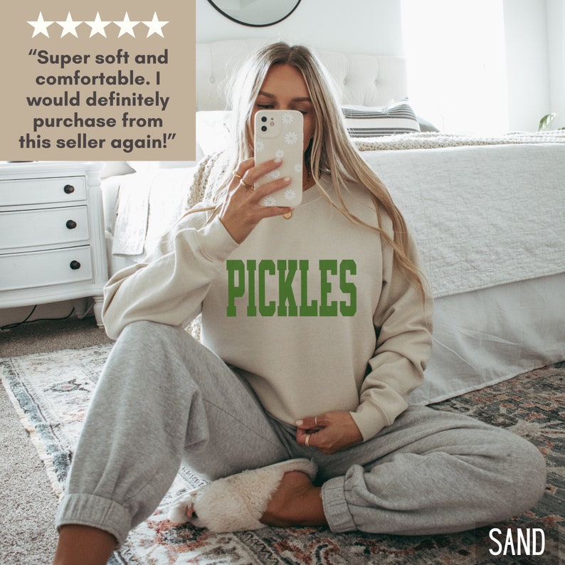 Op de afbeelding: Een licht beige sweatshirt met het woord "PICKLES" erop gedrukt in groene letters. Het sweatshirt wordt gedragen door een persoon die op een tapijt zit.