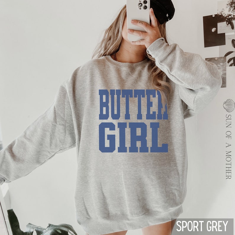 May include: A grey sweatshirt with the text "BUTTER GIRL" printed in blue on the front. The sweatshirt is a relaxed fit and has a crew neck.