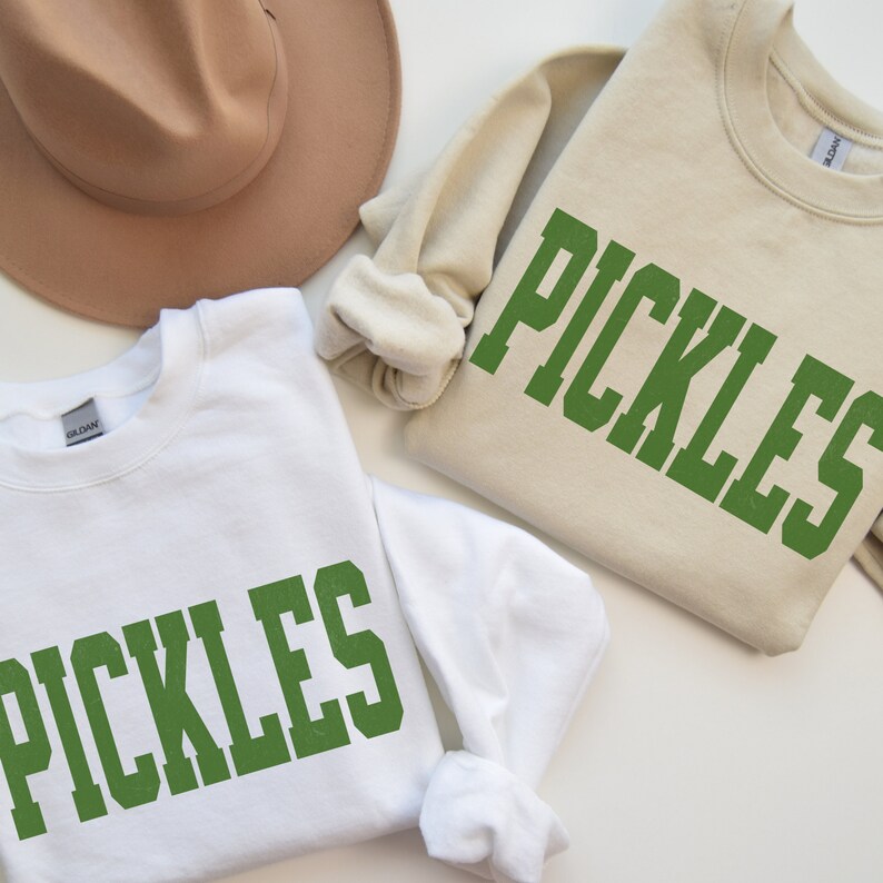 Op de afbeelding: Twee witte sweatshirts met het woord "PICKLES" in het groen op de voorkant bedrukt. De sweatshirts zijn gevouwen en op een wit oppervlak gelegd.