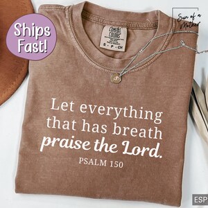May include: Espresso-colored t-shirt with the text "Let everything that has breath praise the Lord. Psalm 150" in white. A silver necklace with a pendant rests on the shirt. The shirt is a Comfort Colors brand.