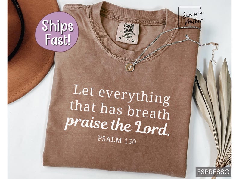 May include: Espresso-colored t-shirt with the text "Let everything that has breath praise the Lord. Psalm 150" in white. A silver necklace with a pendant rests on the shirt. The shirt is a Comfort Colors brand.