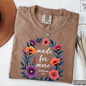 K&ouml;nnte beinhalten: Ein braunes T-Shirt mit einem floralen Kranz-Design und dem Text "made for more Ephesians 2:10".