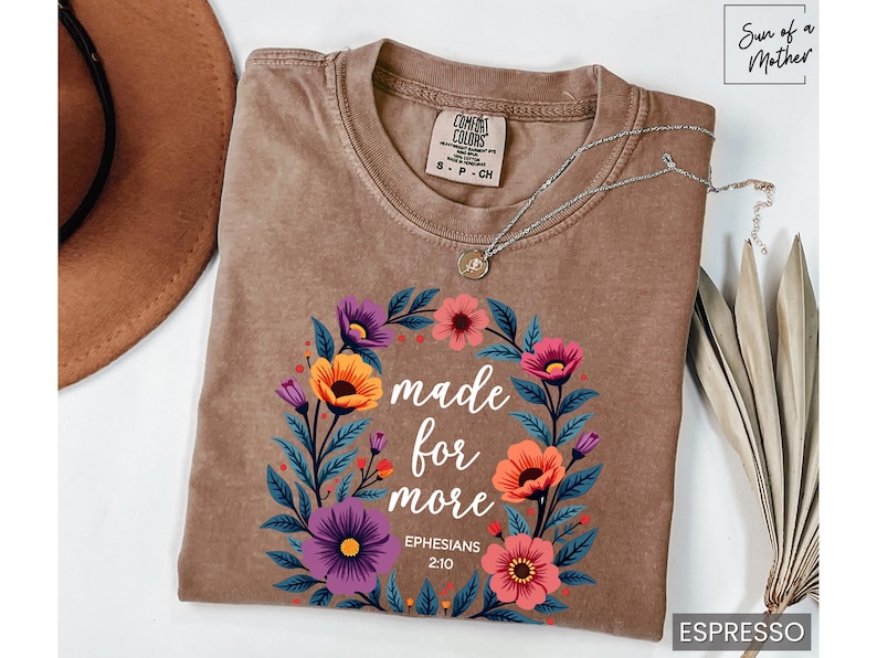 K&ouml;nnte beinhalten: Ein braunes T-Shirt mit einem floralen Kranz-Design und dem Text "made for more Ephesians 2:10".