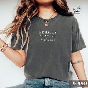 Puede incluir: Una camiseta gris oscuro con el texto "Be Salty Stay Lit" y "Matthew 5:13-14" en letras blancas.